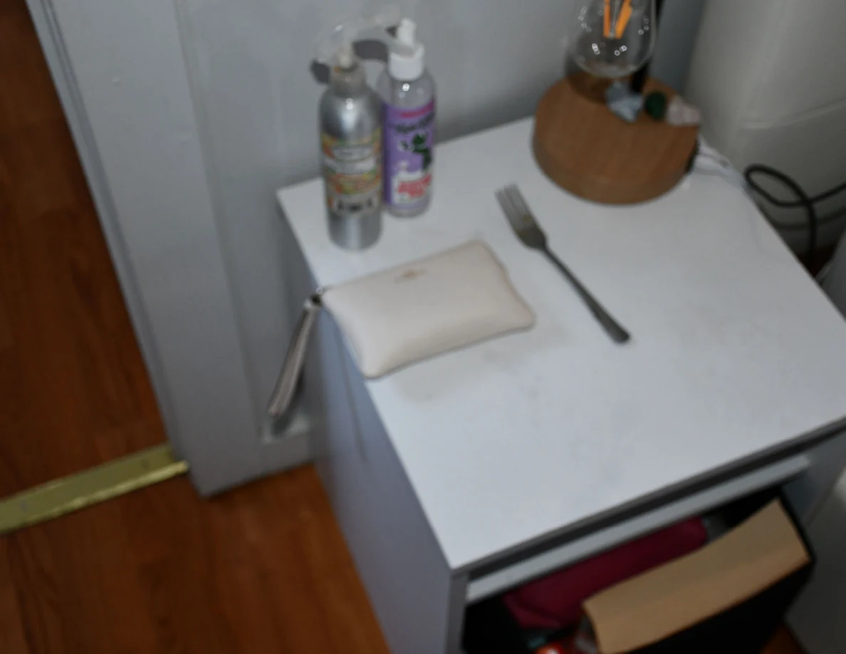 White bedside table holding a small pouch, fork, bottles and lamp above a partly open drawer.
