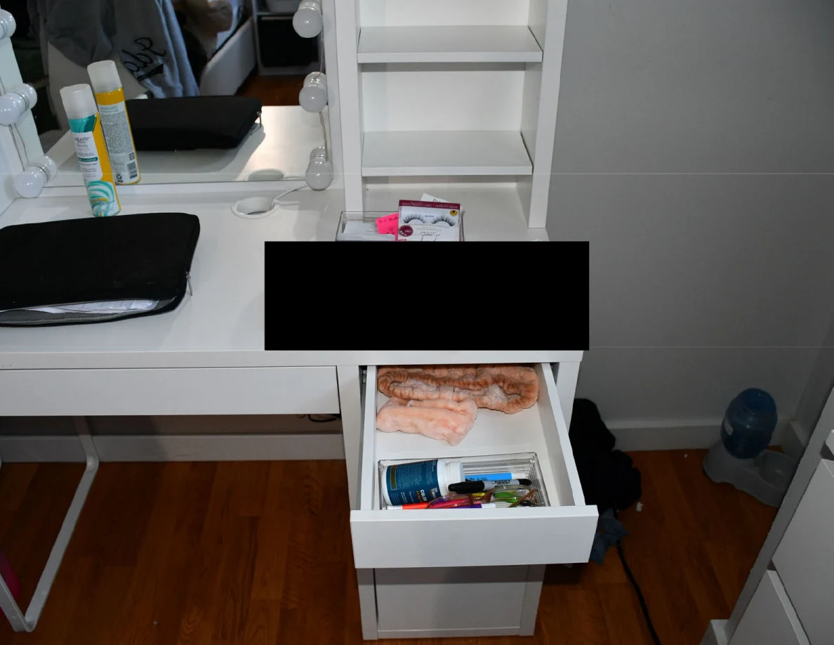 White vanity with an open drawer of small toiletries and objects, and black redactions on the tabletop.