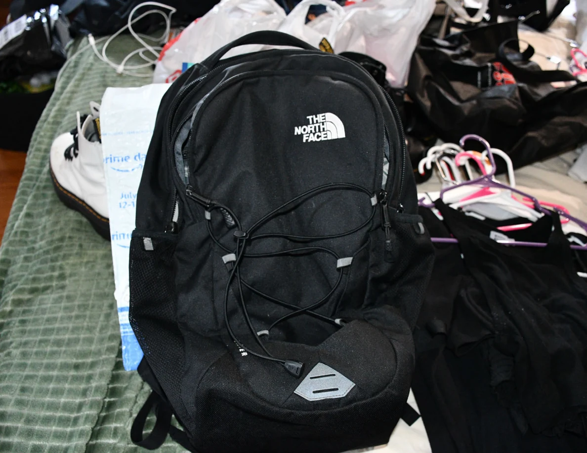 Black backpack resting on a green bed blanket among clothing and hangers.
