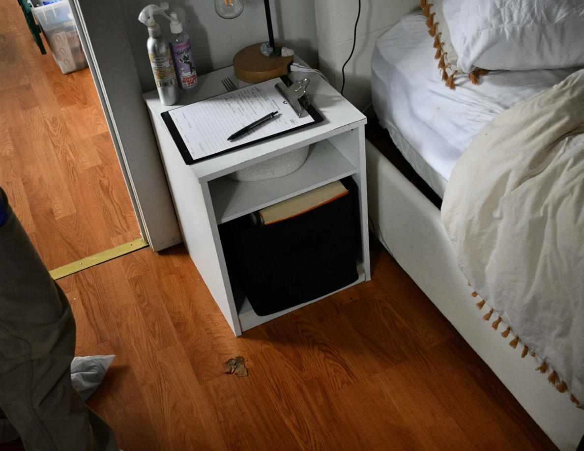 White bedside shelf with clipboard, pen, bottles and lamp above a black storage bin.