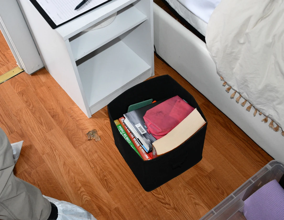 Black fabric storage bin containing books and notebooks on the floor beside a bed.
