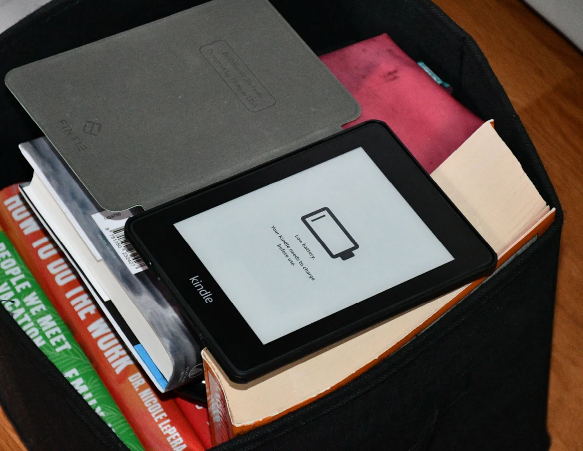 Close view of a Kindle e-reader showing a low-battery symbol atop books in a storage bin.