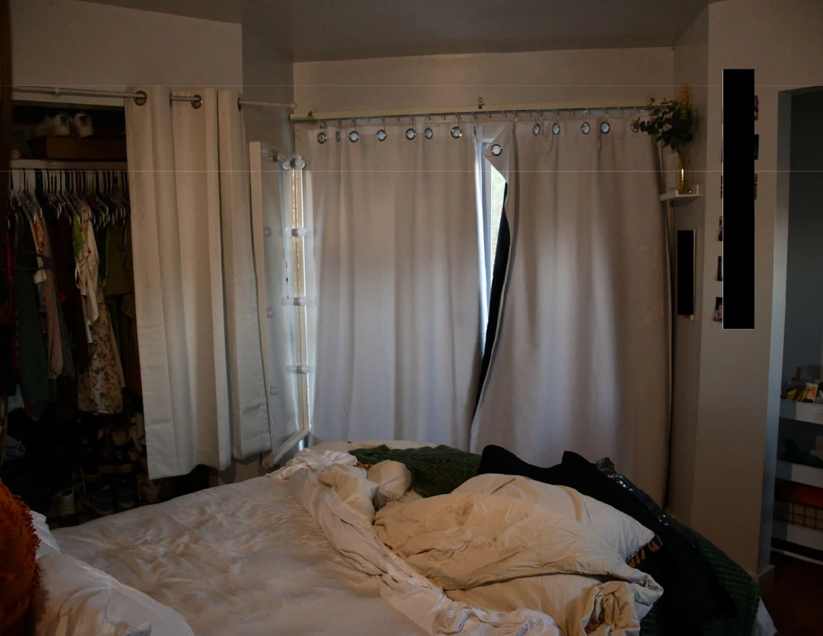 Rumpled bed beside parted curtains, an illuminated mirror and an open clothes closet.