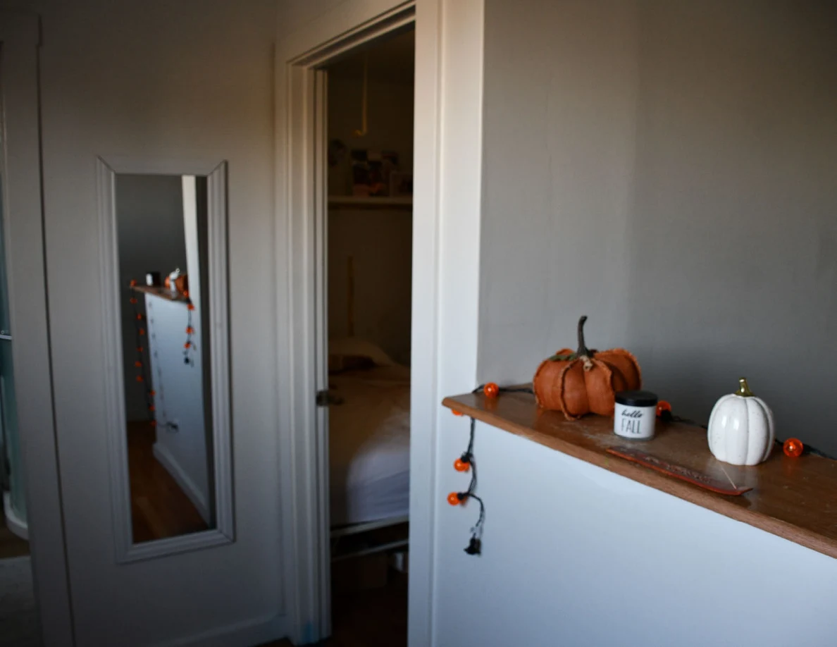 Hall wall with a mirror, doorway and narrow shelf holding pumpkin decorations and string lights.