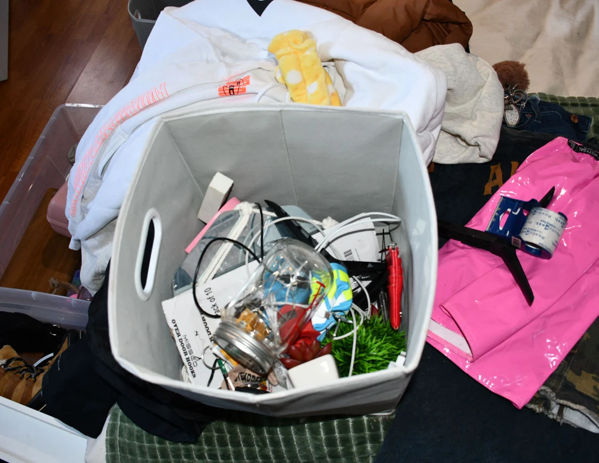 Gray fabric storage bin on a bed containing cords, packaging, glass container and assorted objects.