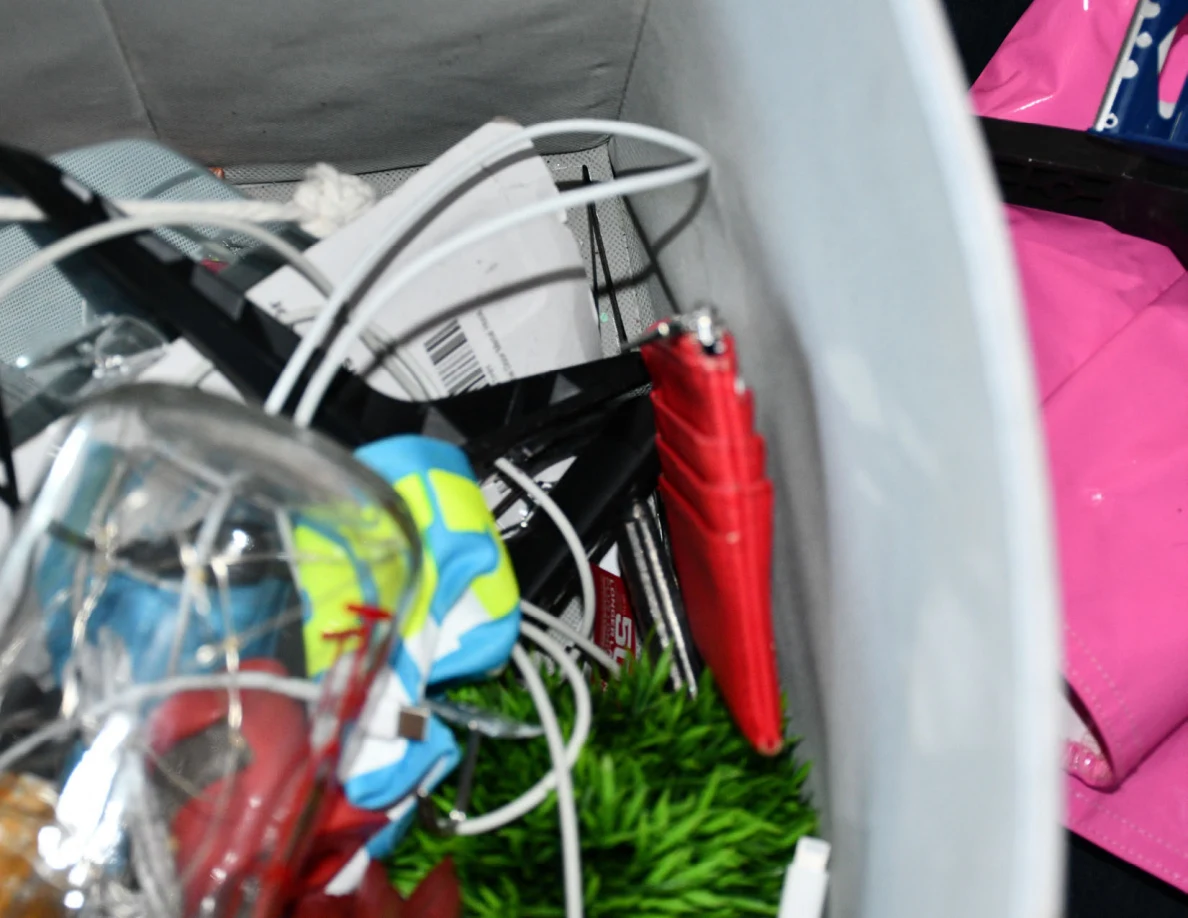 Close view inside a fabric bin with cords, packaging, red wallet and artificial greenery.