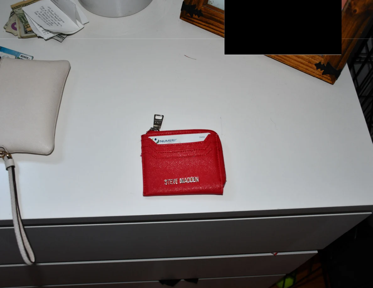 Red zippered card wallet on a white dresser beside a pale wristlet and redacted framed item.
