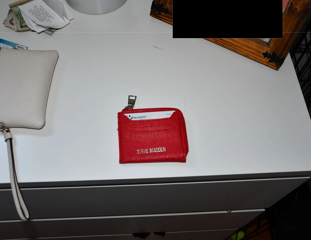 Red card wallet on a dresser beside a wristlet and framed item with redaction.