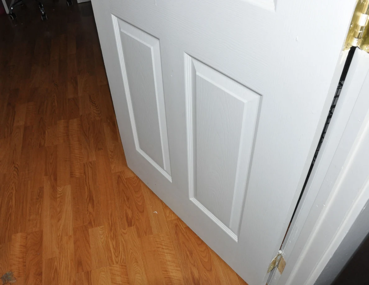 Lower paneled door and brass-colored hinge above wood-pattern flooring.