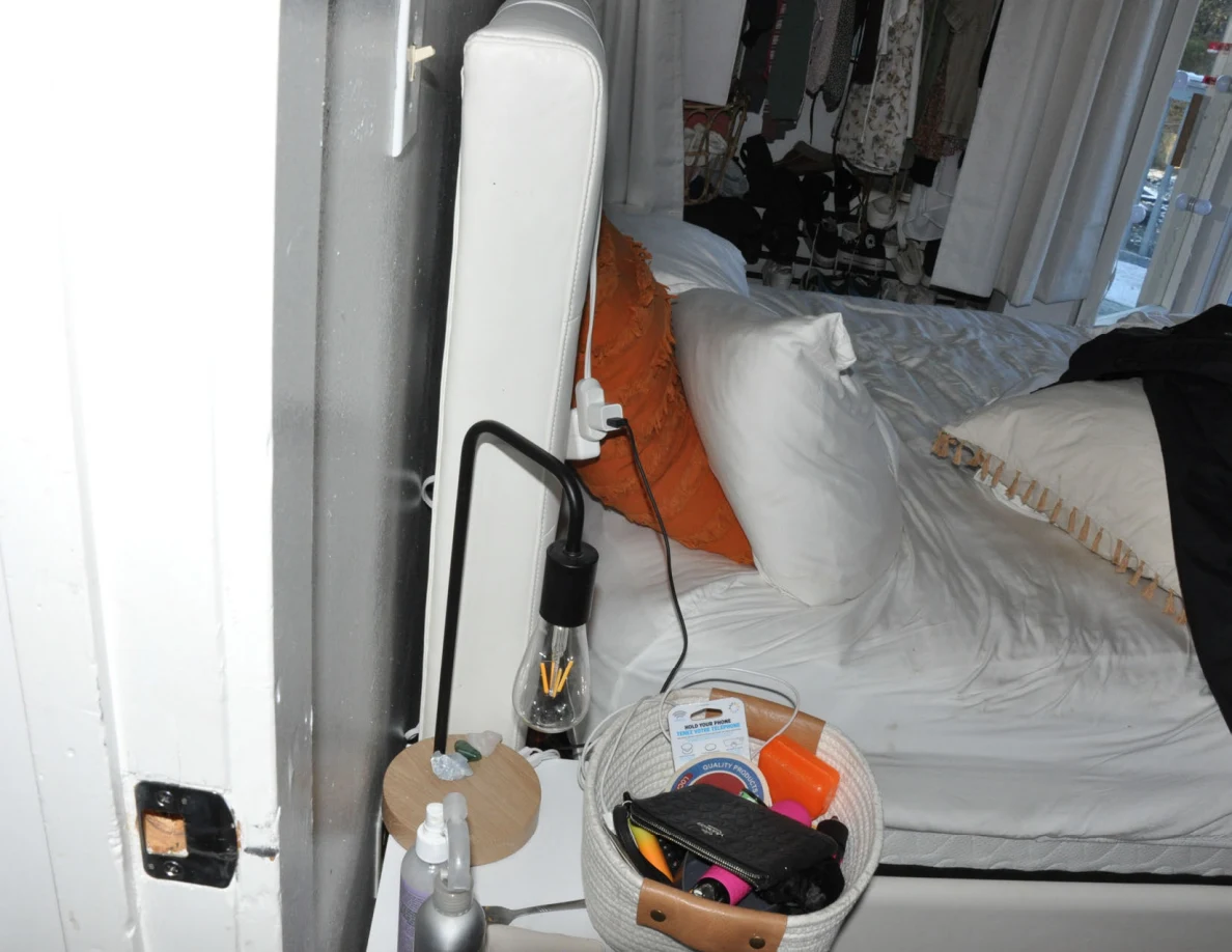 Narrow view beside a bed showing headboard, pillows, lamp and basket on a bedside cabinet.