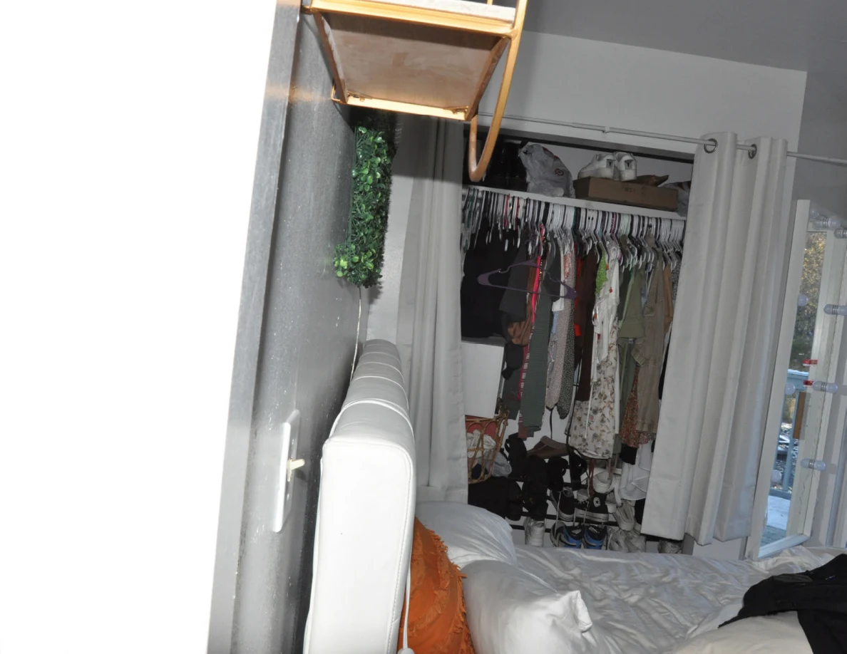 Bed headboard and pillows facing an open closet with hanging clothes.