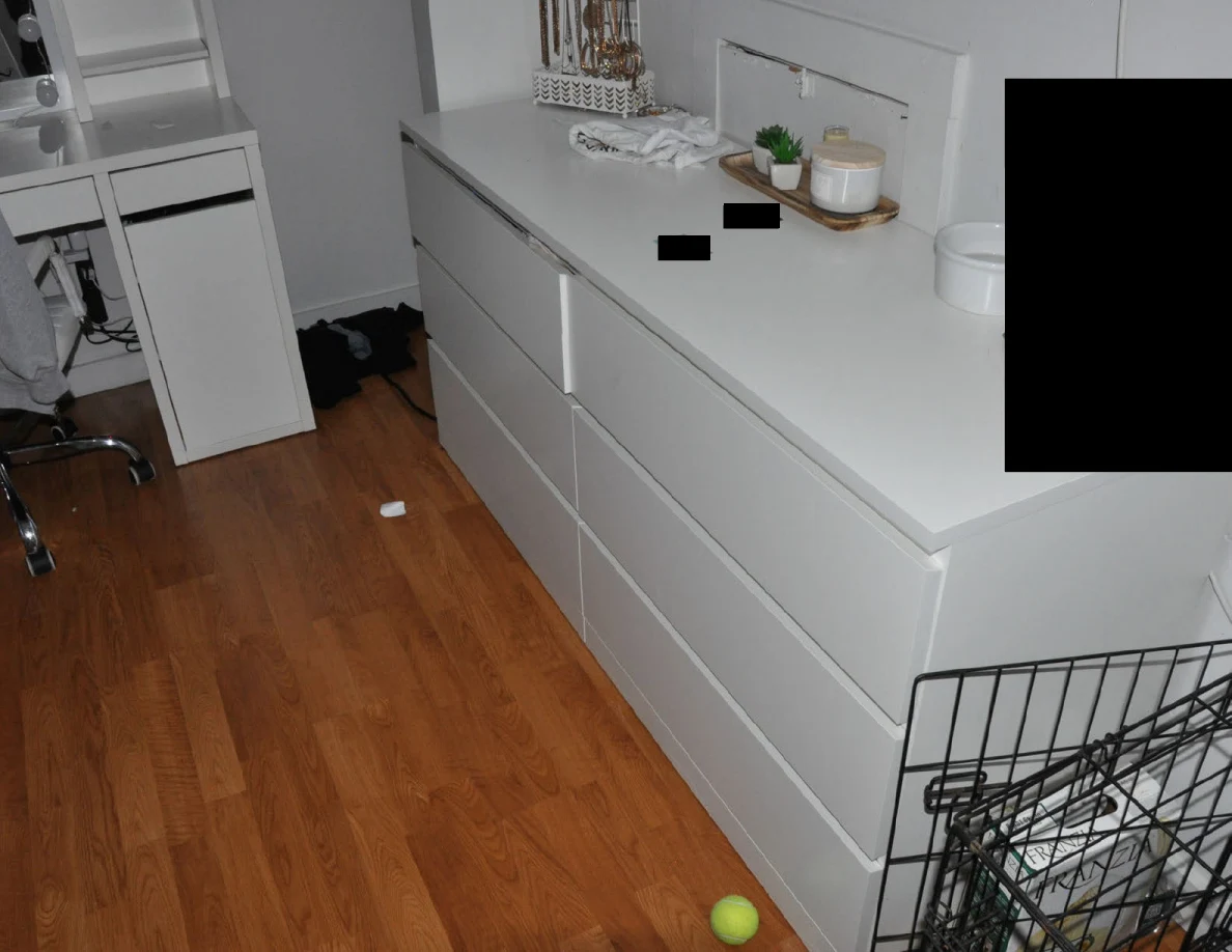 Long white dresser with plants and redacted objects beside a pet crate and tennis ball.