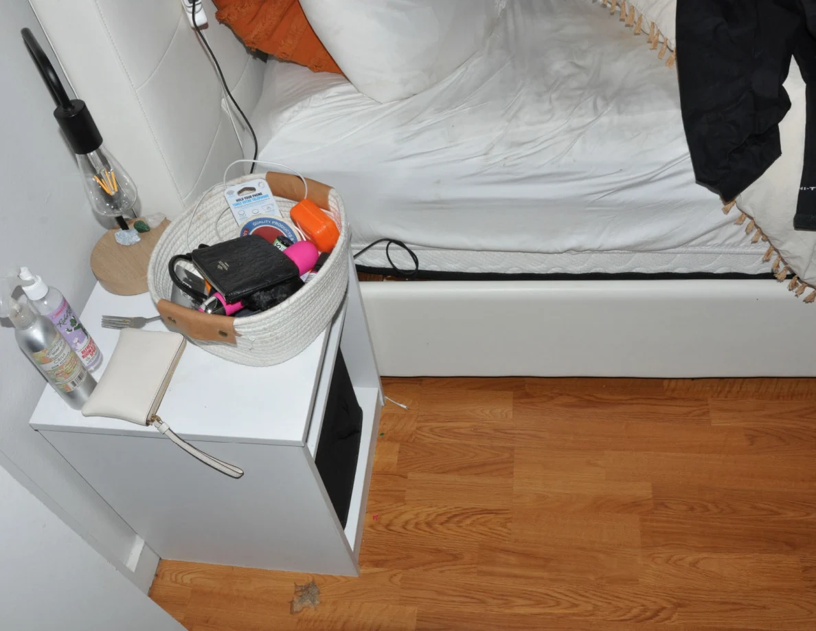 Bedside cabinet holding a woven basket, pouch and lamp beside white bedding.