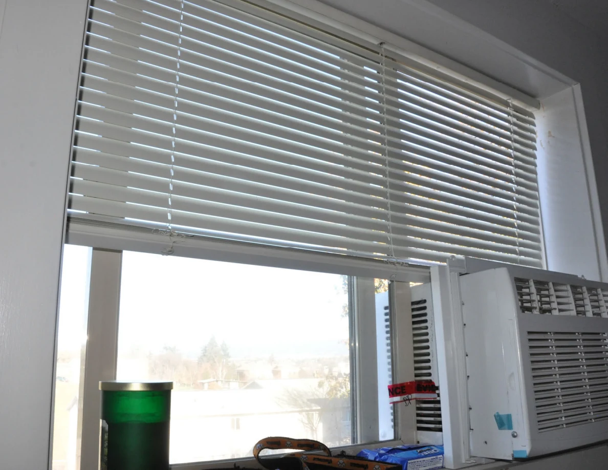 Window blinds above a window air conditioner with a green tumbler on the sill.