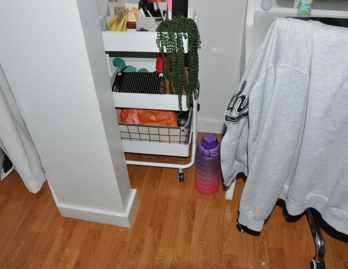 White rolling cart with toiletries and a trailing plant beside a gray sweatshirt and large purple bottle.