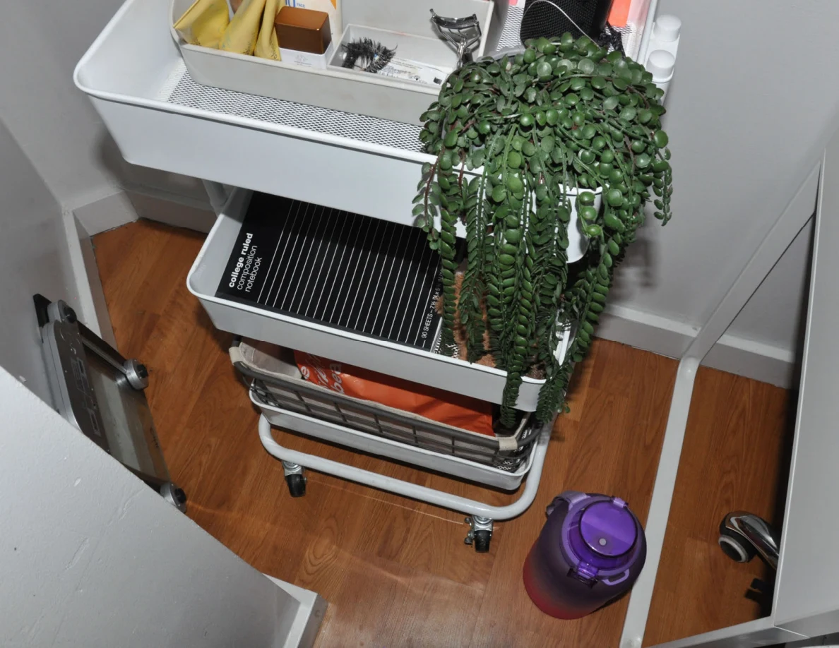 Lower shelves of a rolling cart with notebook, fabric basket and orange shopping bag beside a purple bottle.