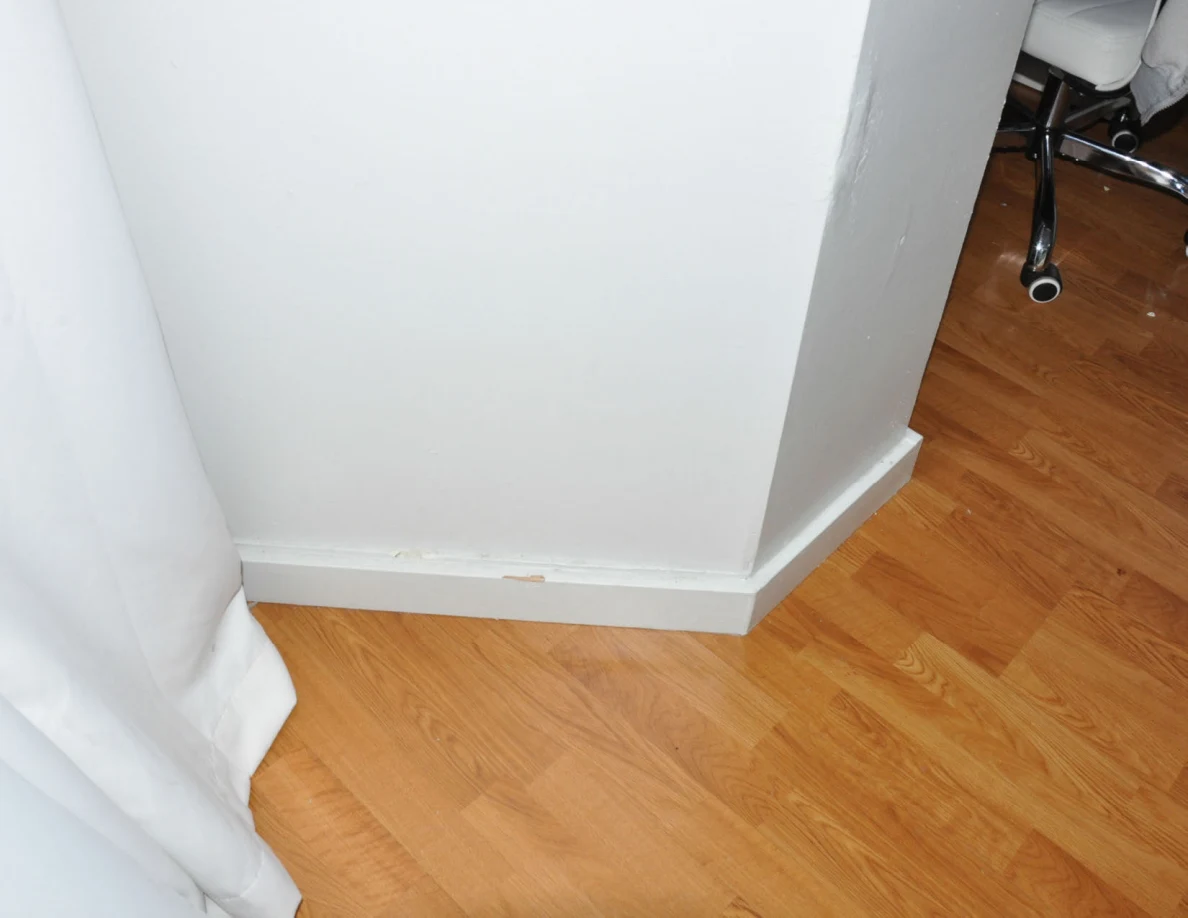 Lower wall corner and white baseboard beside a curtain on wood-pattern flooring.