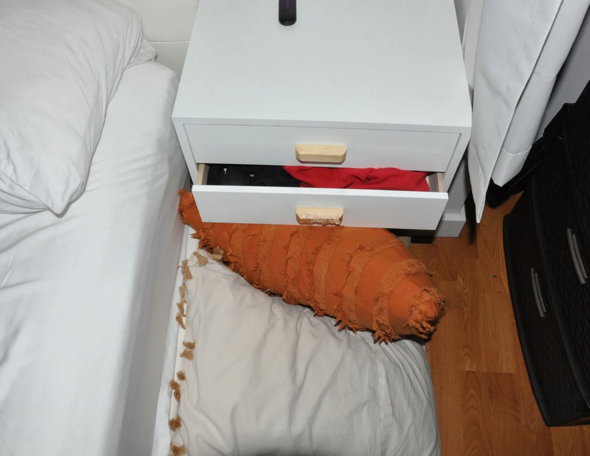 Partly open bedside drawer containing red fabric, with an orange pillow and pale pillow on the floor.