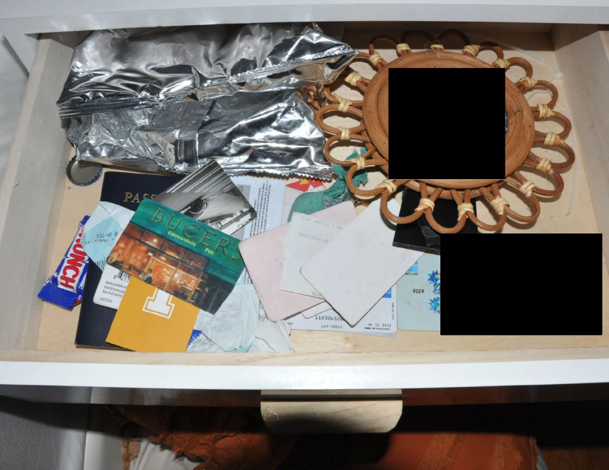 Open bedside drawer with papers, a passport, foil packaging and small decorative mirror, with redactions.