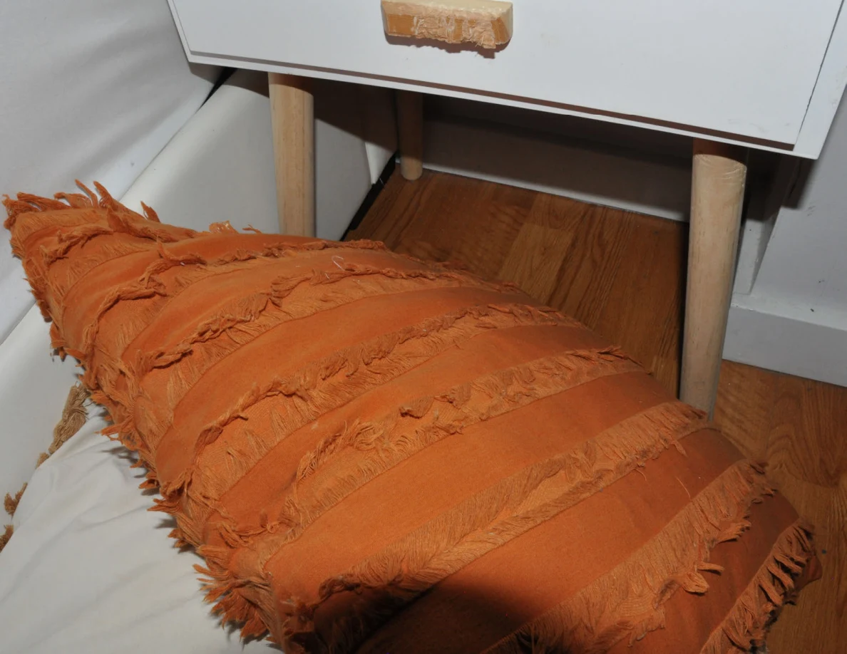 Orange fringed pillow on a pale pillow beneath bedside drawers.