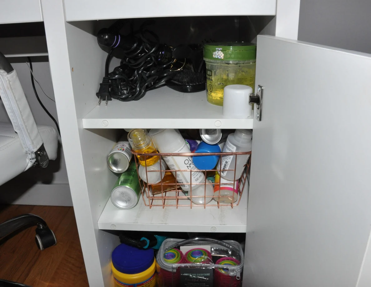 Vanity cupboard shelves holding tangled cords, jars and a wire basket of bottles.