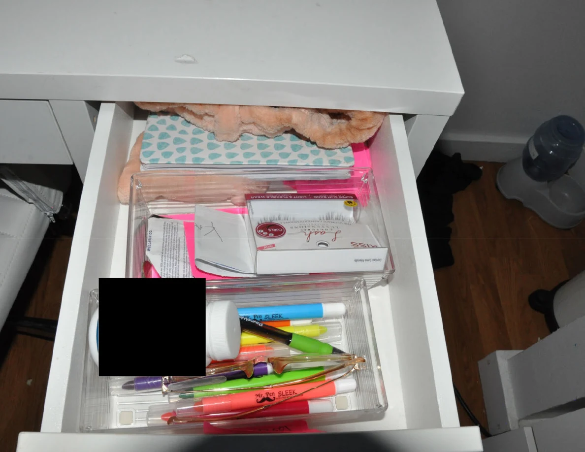 Open vanity drawer holding pens, cosmetic packaging and small notebooks with redacted items.