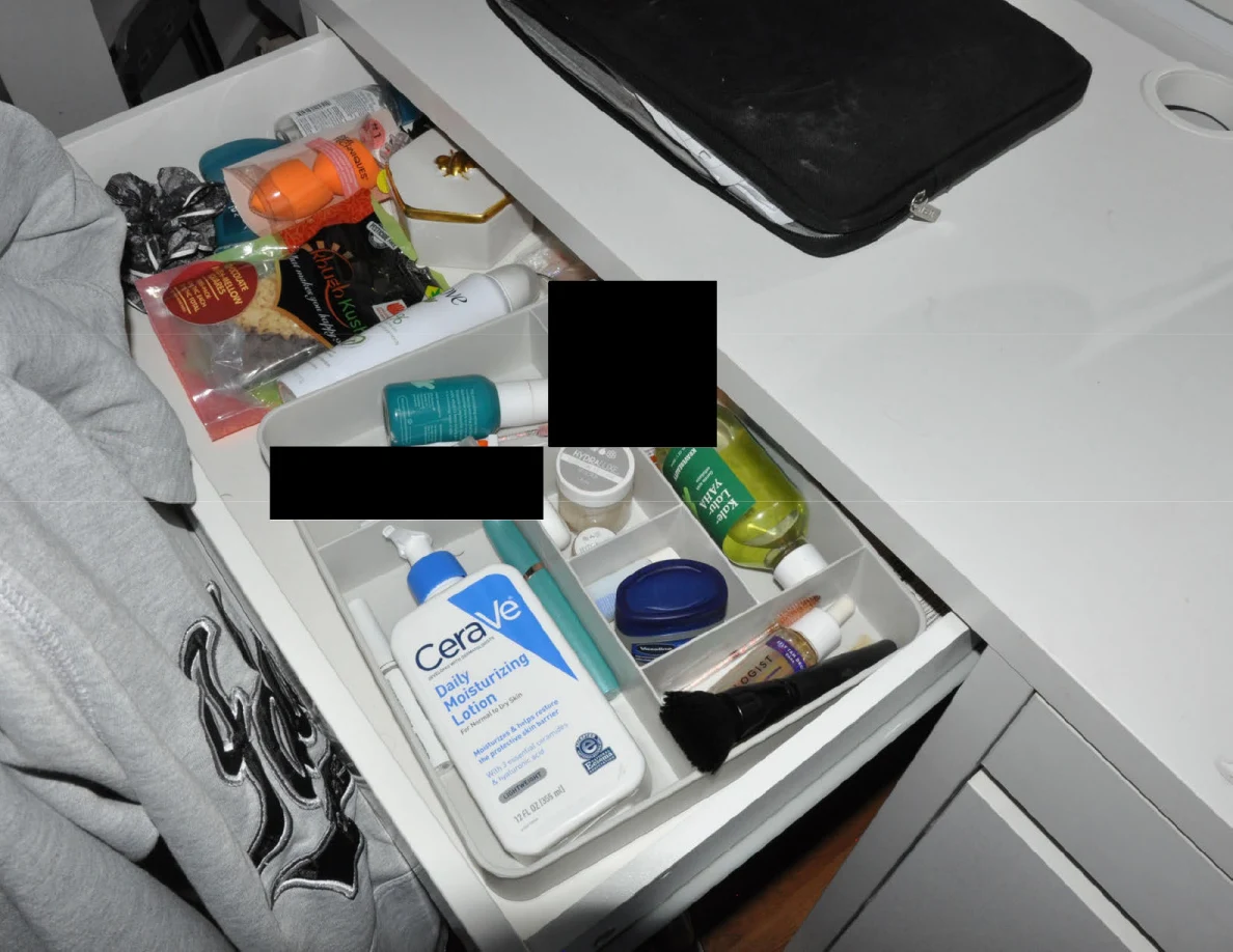 Wide vanity drawer containing lotion, toiletries and small objects with black redactions.