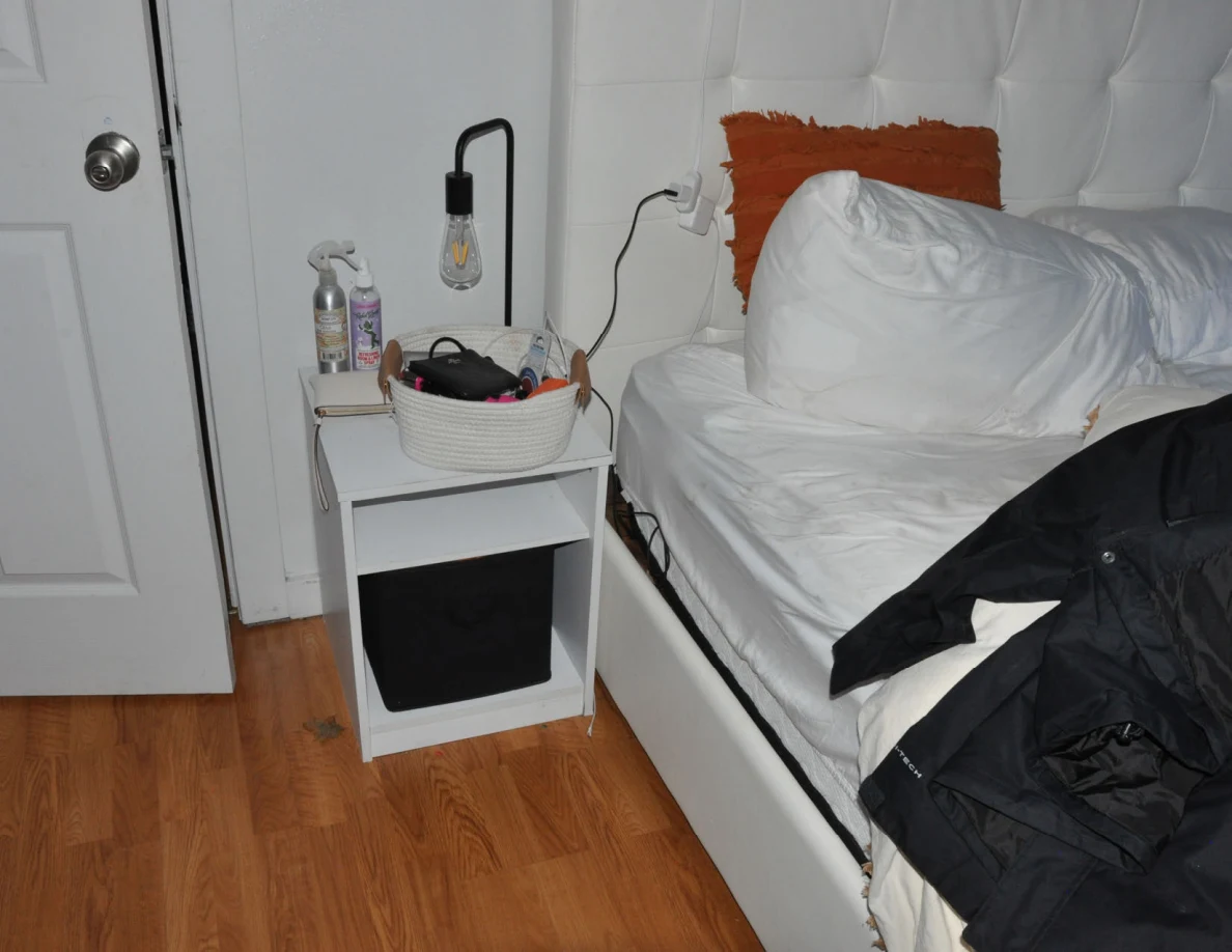 White bedside shelf and lamp next to pillows and a dark garment on a bed.