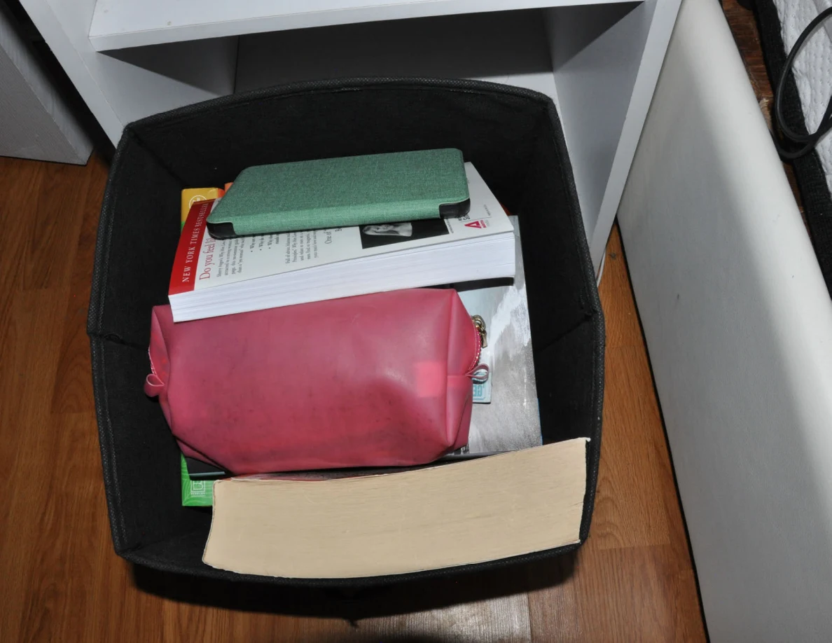 Black fabric bin holding books, a green-covered item and pink pouch beside a bed.
