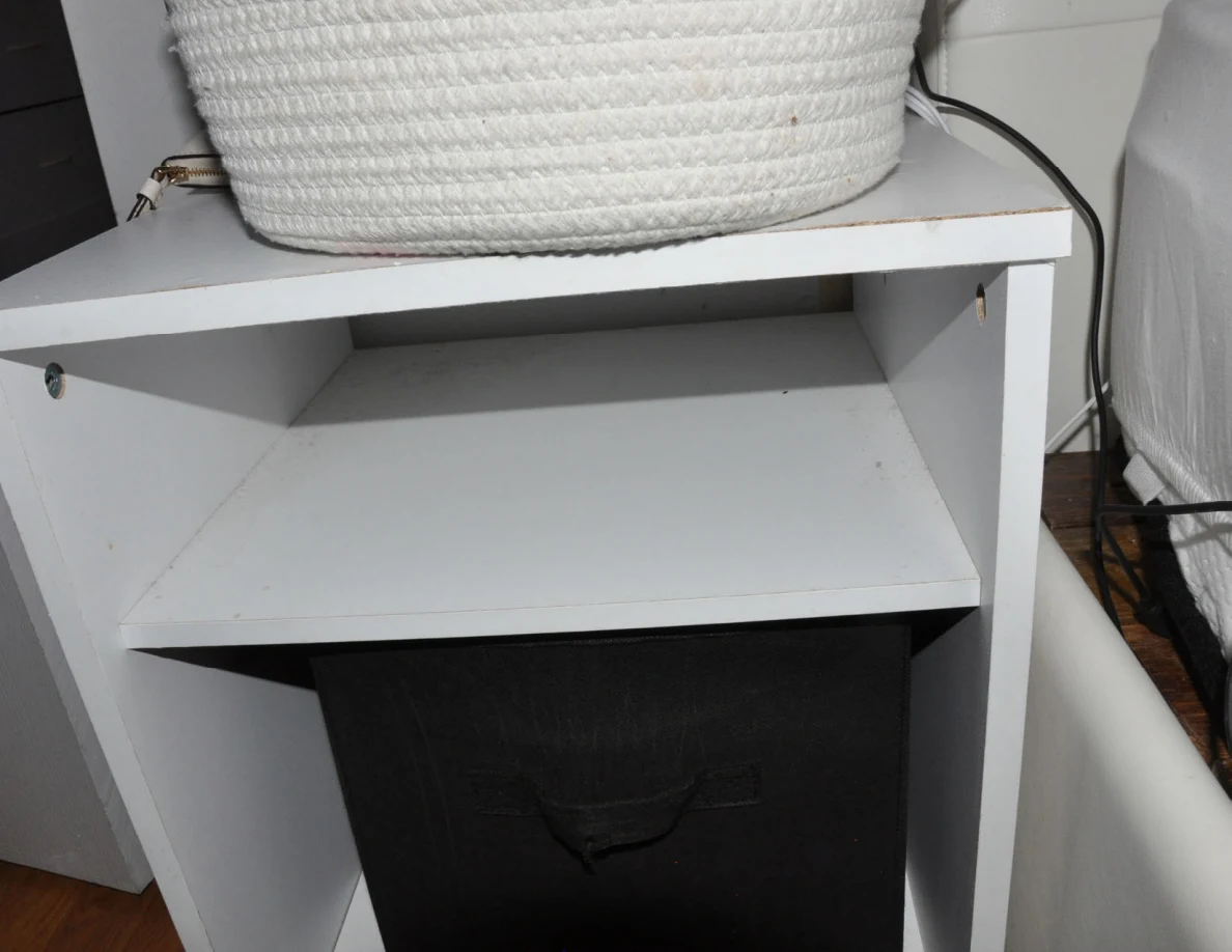 White bedside shelving with a woven basket on top and black fabric bin below.