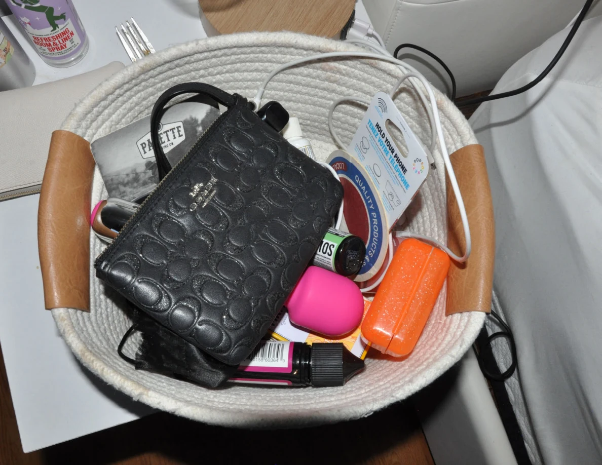 Woven bedside basket containing a black patterned wallet, cords and small cases and toiletries.