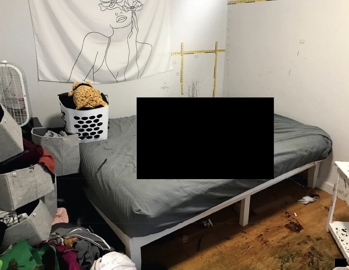 Gray-covered bed beneath a figure wall hanging beside a laundry basket and measuring tape, with a large redaction.
