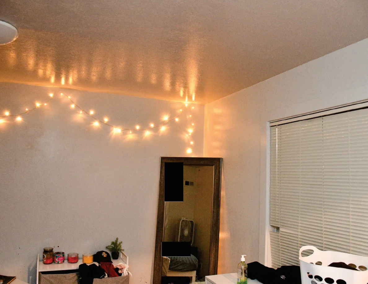 Glowing string lights across a bedroom ceiling corner above a tall mirror and window blinds.