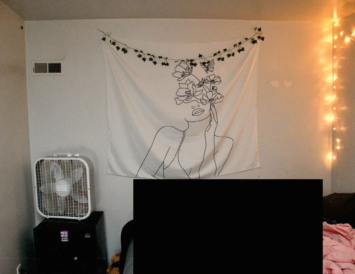 Figure wall hanging above a box fan on a black cabinet beside a heavily redacted bed.