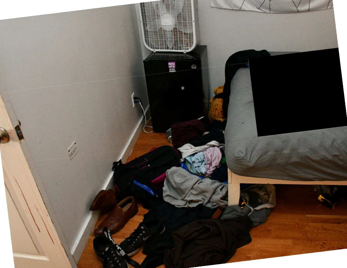 Angled view of shoes and clothing on the floor beside a bed and box fan, with redaction.