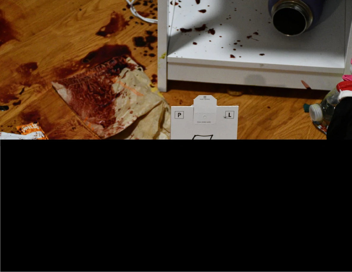 Reddish-brown marks on a wood-pattern floor and crumpled paper beside a white shelf, with much of the image redacted.