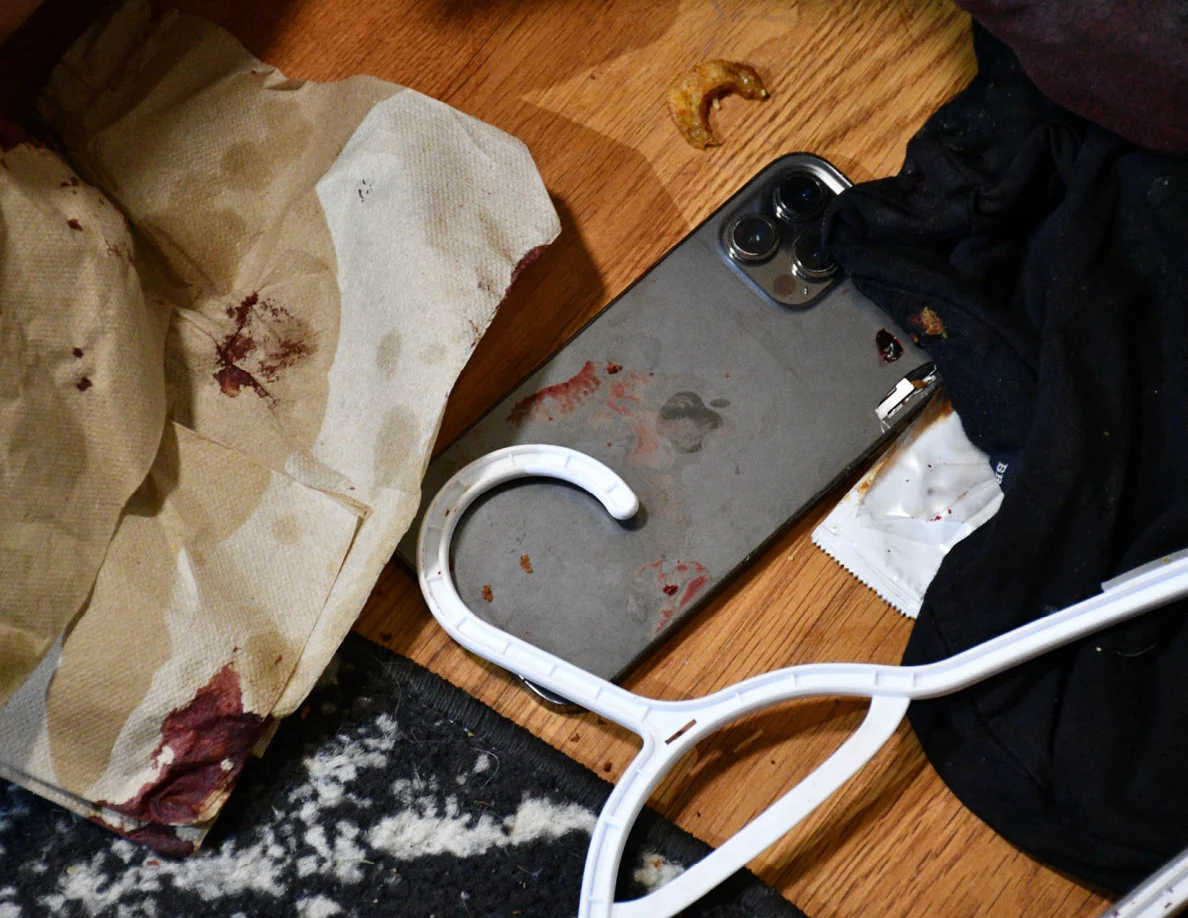 Phone lying among a white clothes hanger, dark fabric and marked paper on a wood-pattern floor.