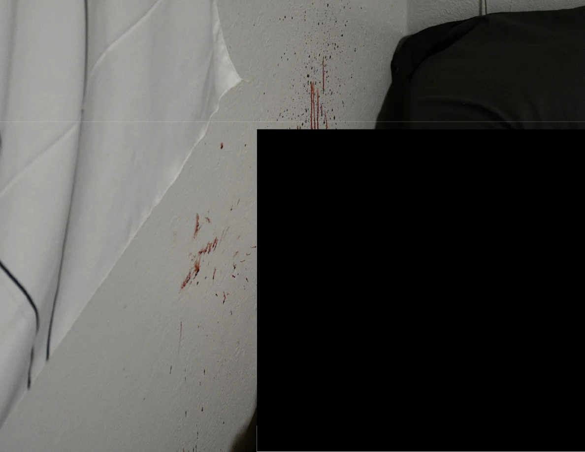 Reddish streaks and spots on a pale wall beside dark bedding and a large redaction.