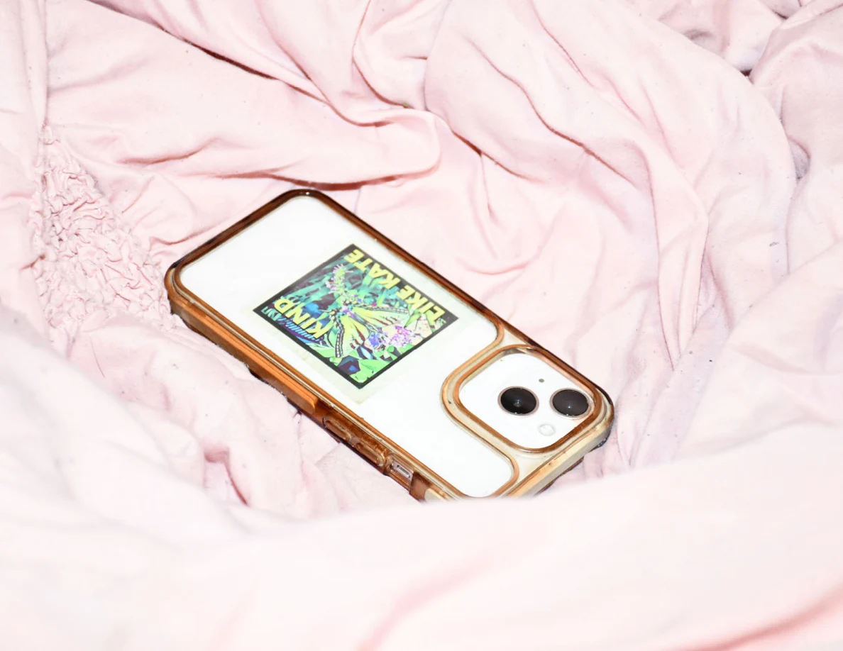 Close view of a white phone in a clear amber-edged case with a colorful sticker on pink bedding.