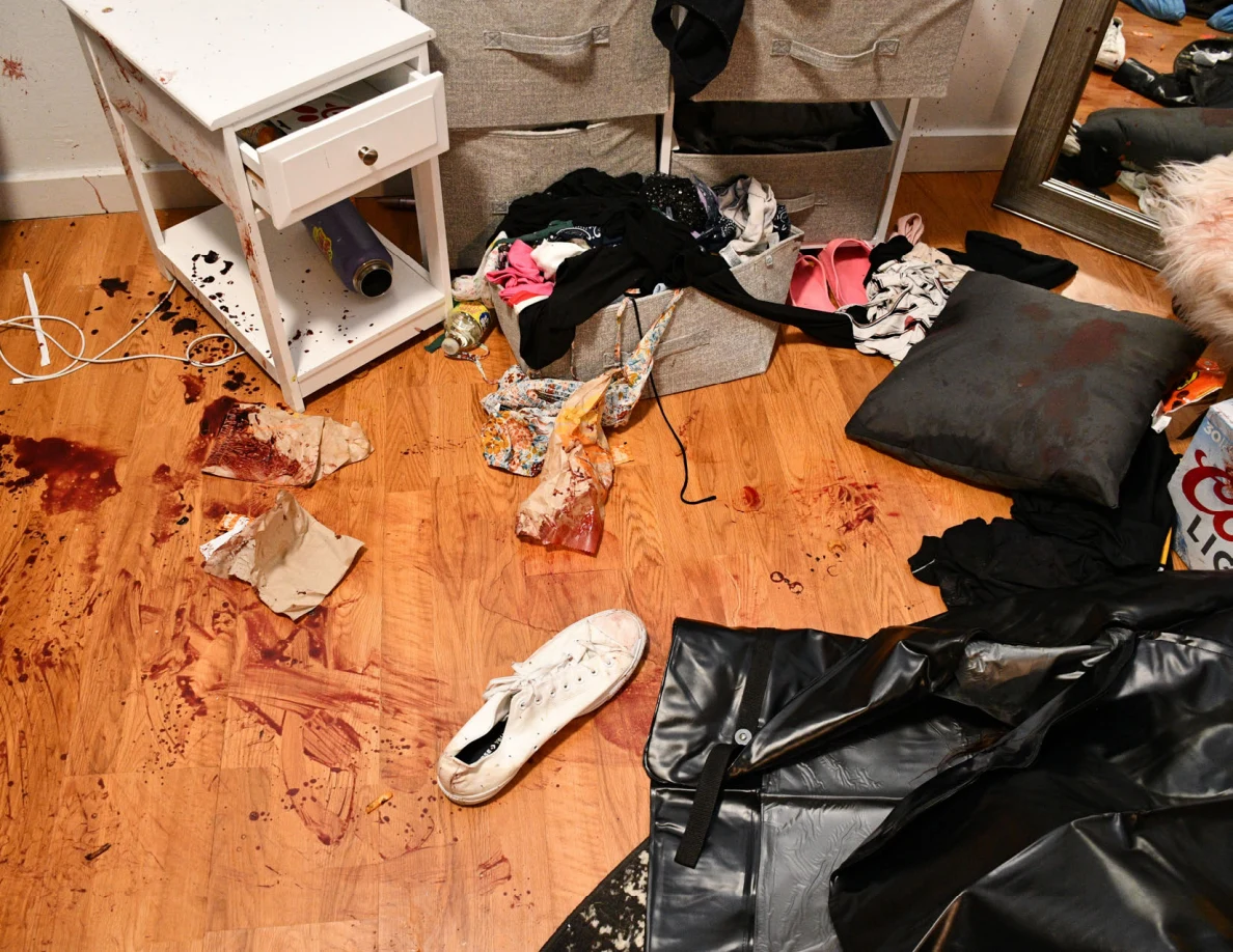 Bedroom floor with reddish-brown marks, crumpled paper, white shoe, scattered clothing and black sheeting beside a small white cabinet.