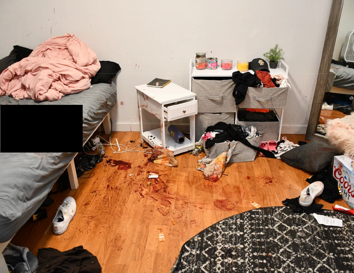Bed edge, white bedside cabinet and fabric drawers above a floor with reddish-brown marks and scattered clothing.