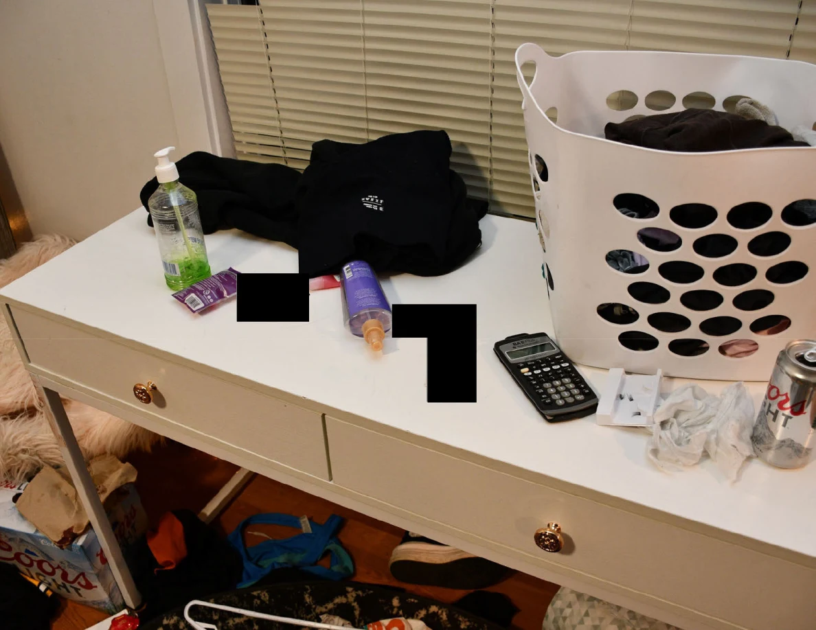 White desk with laundry basket, calculator, toiletry bottles and dark clothing, with redacted items.