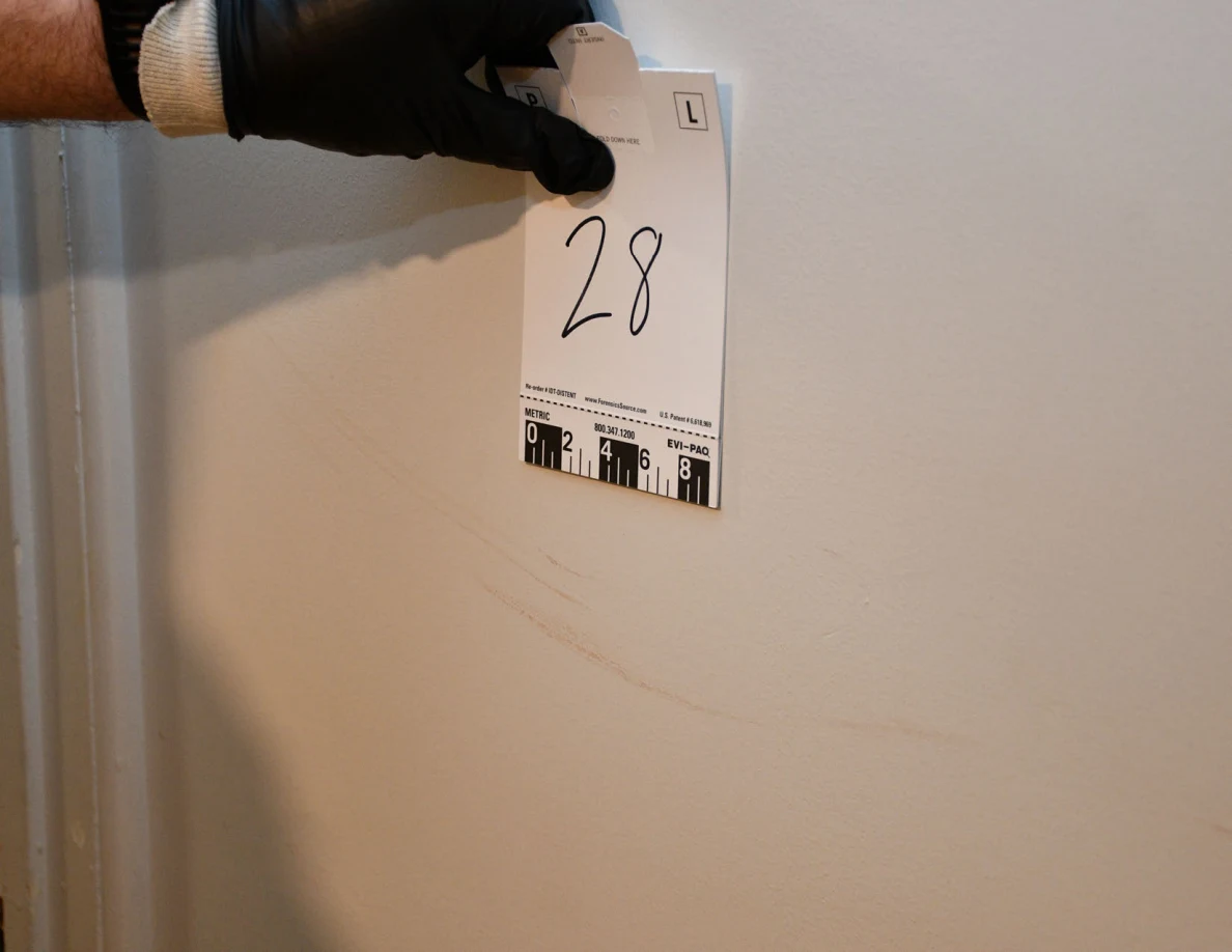Black-gloved hand holding scale card 28 beside a faint curved mark on a pale wall.