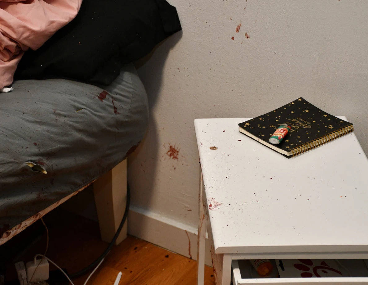 Notebook and small cylindrical item on a white bedside cabinet beside reddish wall spots.