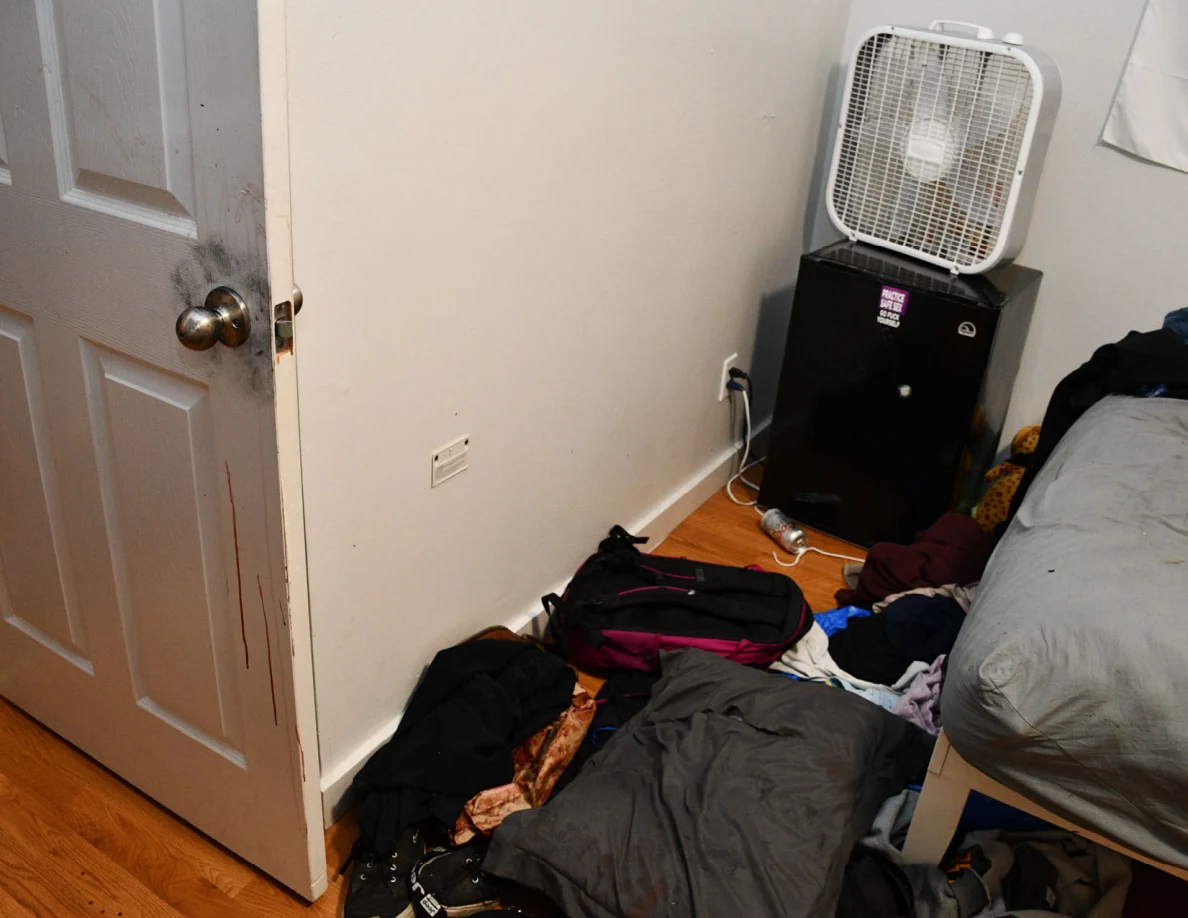 Open white door beside a box fan on a dark cabinet and clothing near a bed.