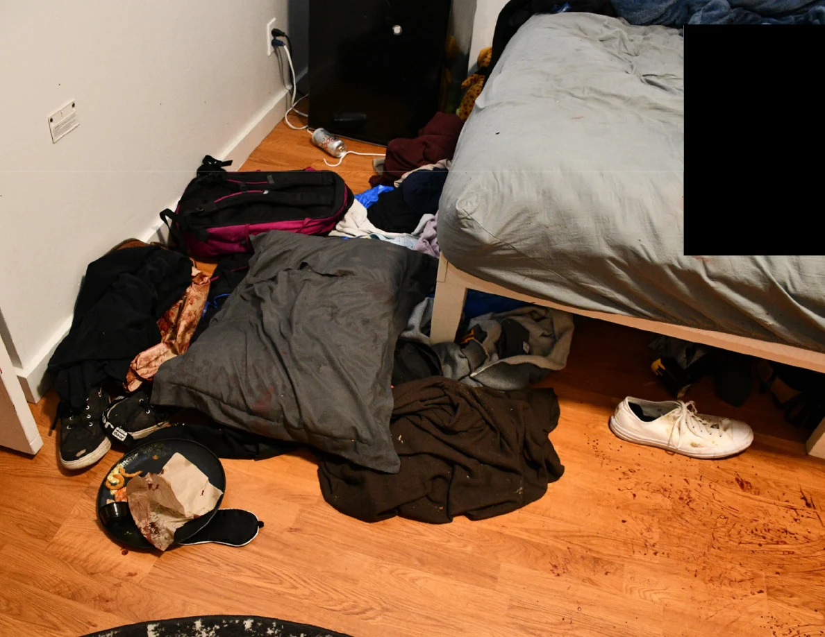 Clothing, bags and a white shoe beside a bed, with a dark round tray and food paper.