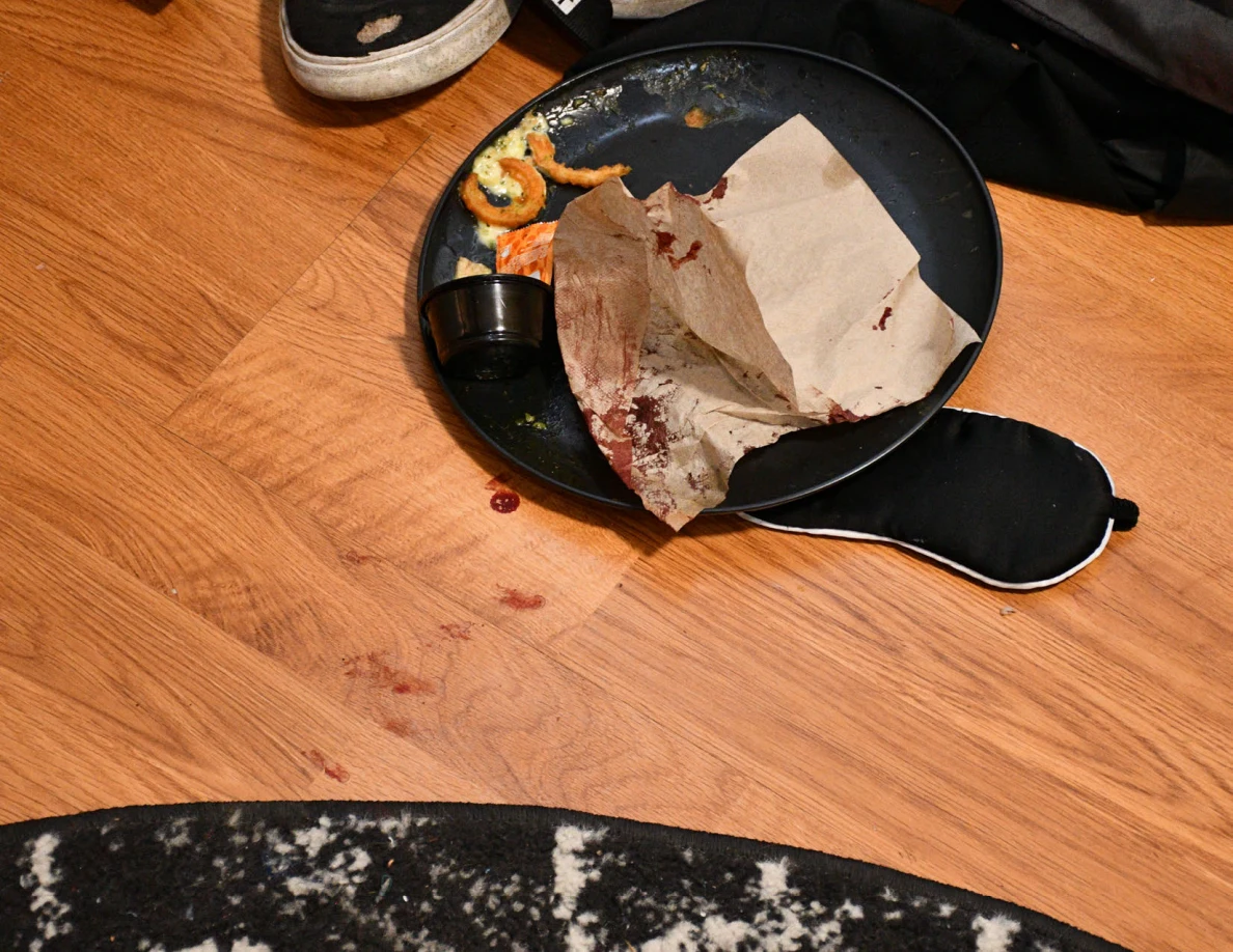 Dark round tray with food remnants, a paper bag, small dark cup and eye mask beside reddish floor marks.
