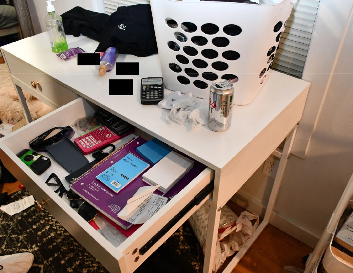 Open white desk drawer containing notebooks, calculators and paper beside a laundry basket.