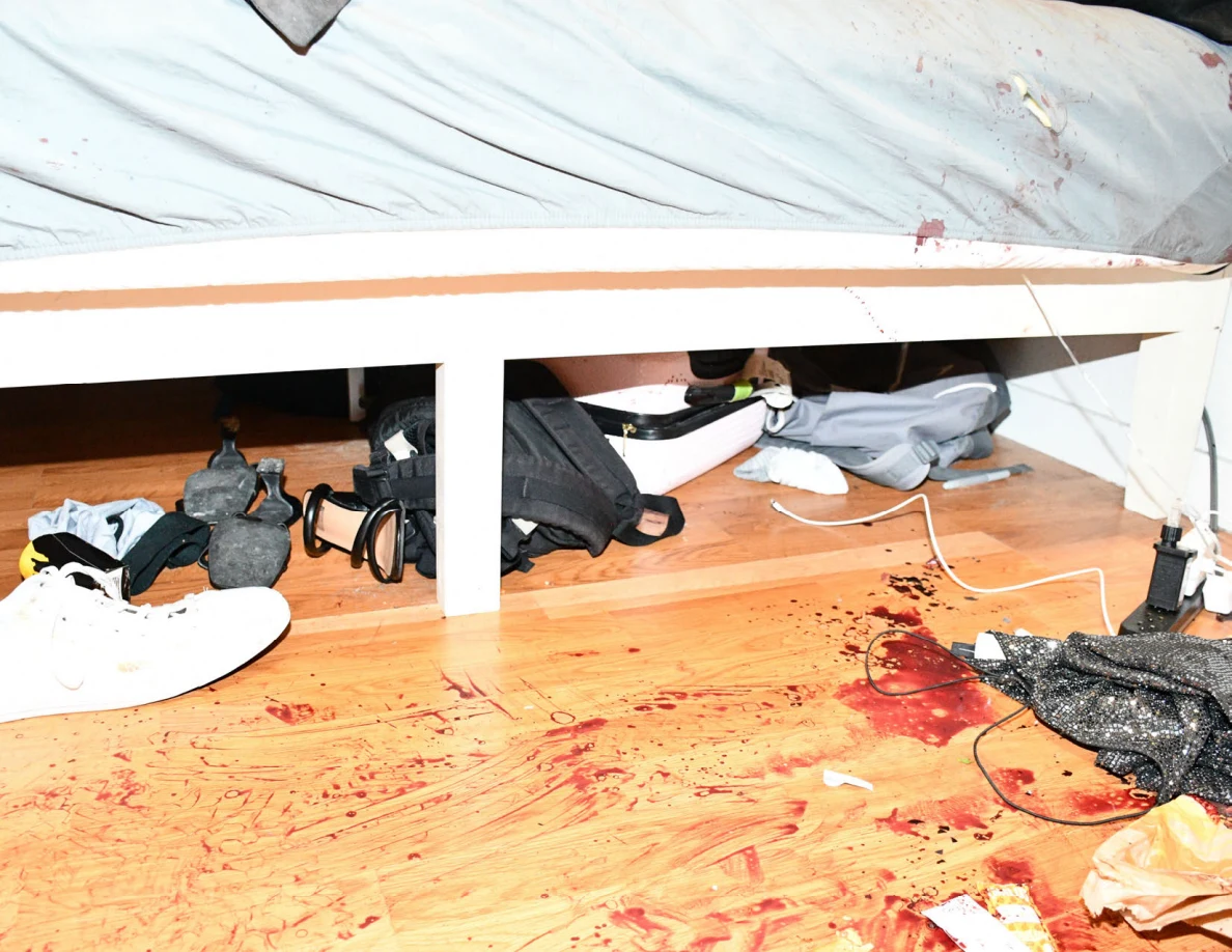 Bed frame and gray sheet above reddish floor marks, cables, footwear and bags.