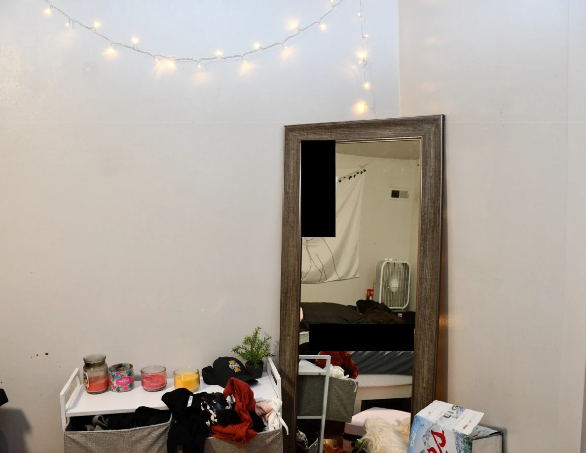 Tall framed mirror with a black redaction above fabric drawers, candles and clothing beneath string lights.